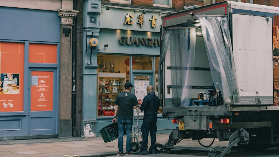 Two men are engaged in a home relocation task outside a shop named 'Guanghua' in Kentish Town. One man, wearing a black cap and dark clothing, holds a black plastic crate, while the other, dressed in a navy jacket, is standing beside a small wheeled sack trolley. They appear to be preparing to load or unload items from a large, open-sided delivery truck parked adjacent to the shop. The truck's cargo area, visible in the image, contains packed boxes and packing materials, with clear plastic sheets covering part of the interior. The shopfront features a glass display window with various items inside, and signage with gold Chinese characters above the entrance. The scene is set on a paved sidewalk, with the building's facade and neighboring structures visible in the background, capturing a typical urban environment associated with moving and packing activities conducted by companies like Man with Van Kentish Town during a furniture transport or home relocation process.