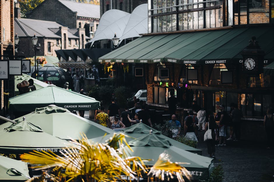 A busy outdoor scene in Camden Town showing several green umbrellas with 'Camden Market' branding, shading outdoor seating areas. Behind the umbrellas, there are groups of people walking and standing on the pavement outside a building with a green awning displaying 'Camden Market' and a clock mounted on the wall. The building has brick walls and large windows, with some hanging flower baskets and a glass-domed structure visible on the rooftops. In the foreground, we see various cardboard boxes and packing materials, possibly prepared for a home relocation or furniture transport. The loading area is adjacent to the pavement, where a man with a trolley might be carrying boxes toward a nearby van, which is partially visible behind the crowd. The scene is well-lit with natural daylight, capturing the movement and activity typical of a commercial and crowded urban environment. Occasionally, Man with Van Kentish Town might assist such moving logistics involving packing and transporting household items through busy streets like these, supporting Camden council removal permits.