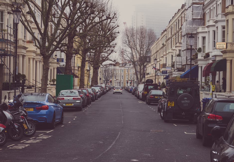 A view of a residential street in Kentish Town with parked cars lining both sides of the road and a woman walking alongside a cyclist on the pavement. The street features a mixture of small brick buildings and trees providing greenery. In the background, a large historic church with a prominent steeple and Gothic architectural details stands against a partly cloudy sky. Street lamps and traffic signage are visible along the pavement. The scene captures a typical urban setting suitable for house removals or furniture transport, with no visible furniture or moving equipment present in the image. This setting reflects the typical environment in which a professional removals service like Man with Van Kentish Town might operate during a home relocation, emphasizing the importance of planning routes and parking for efficient furniture transport along this area near Fortess Road.