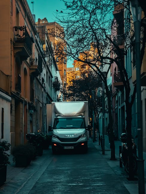 A white Iveco moving van with a large cargo area parked in a narrow urban street lined with multi-story residential buildings and leafless trees. The van's cargo area is covered with a grey protective sheet, indicating it is used for home relocation or furniture transport. The street has a paved sidewalk with black bollards and a bicycle locked to one of them. The surrounding buildings feature balconies, windows, and typical city architecture, with warm sunlight casting shadows and highlighting the scene. The environment suggests a routine loading process for house removals, consistent with professional moving services offered by Man with Van Kentish Town in the context of packing and moving in the Kentish Town area.