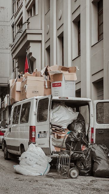 A white Iveco moving van with a large cargo area parked in a narrow urban street lined with multi-story residential buildings and leafless trees. The van's cargo area is covered with a grey protective sheet, indicating it is used for home relocation or furniture transport. The street has a paved sidewalk with black bollards and a bicycle locked to one of them. The surrounding buildings feature balconies, windows, and typical city architecture, with warm sunlight casting shadows and highlighting the scene. The environment suggests a routine loading process for house removals, consistent with professional moving services offered by Man with Van Kentish Town in the context of packing and moving in the Kentish Town area.