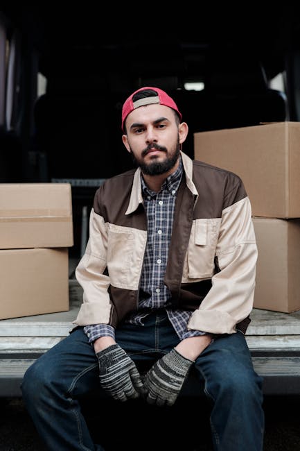A young man with a beard, wearing a red baseball cap backward, a brown and beige utility jacket over a checkered shirt, sits on the edge of an open van cargo area during a home relocation. Surrounding him are several neatly stacked cardboard boxes of various sizes, some secured with plastic wrapping, ready for moving. His hands rest on his knees, which are covered by dark jeans, and he wears grey and black striped gloves, indicating handling and packing tasks. The van's interior is visible behind him, with a dark back panel, and the setting appears to be on a residential street, with natural daylight illuminating the scene. The image reflects a professional furniture transport and packing process, in line with moving services provided by Man with Van Kentish Town, supporting the seamless logistics of house removals and home relocation projects.