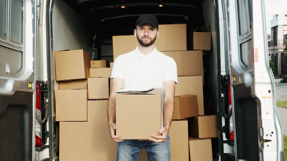 A man with a beard and wearing a black cap, white polo shirt, and blue jeans stands inside the open rear of a large moving van, holding a cardboard box with a clipboard resting on top. The van is loaded with numerous cardboard boxes of various sizes, some stacked on top of each other and others placed side by side, indicating a home relocation process. The interior of the van is spacious, with boxes arranged neatly for transport. The background shows a suburban street with residential houses and greenery, suggesting the moving activity is taking place outdoors. The lighting is natural, highlighting the details of the boxes and the man’s facial expression as he looks towards the camera. This scene captures an essential moment during furniture transport and packing for a house move, with professional removal services provided by Man with Van Kentish Town, supporting efficient and organized moving logistics.