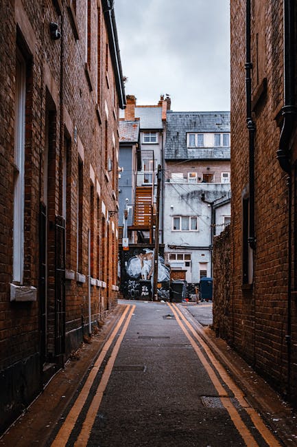 A narrow alleyway between two brick buildings in Kentish Town, showing the back entrances and fire escapes of residential properties. The ground is paved with asphalt and marked with yellow double lines along the edges. At the end of the alley, there is a view of a small courtyard area with stacked cardboard boxes, plastic-wrapped furniture, and large package bags, indicating preparations for a home relocation or furniture transport. The scene is illuminated by natural daylight, with some areas of shadow cast by the surrounding structures. A man with a van, part of the Man with Van Kentish Town service, is likely involved in loading or unloading items as part of a professional moving or packing and moving operation, suitable for house removals in the narrow streets of KENTISH TOWN.