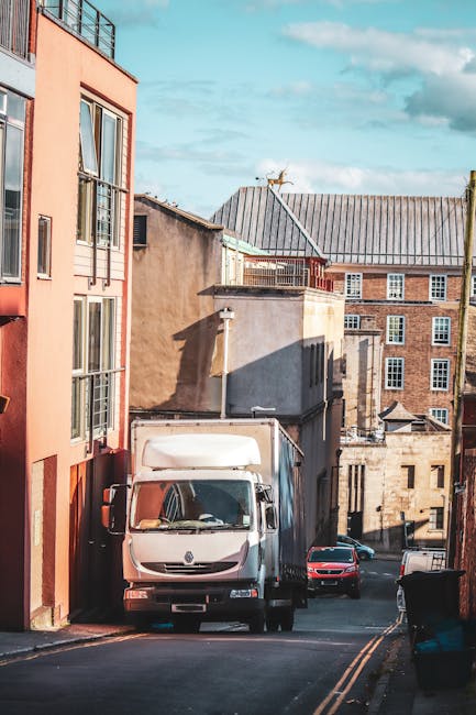 A narrow alleyway between two brick buildings in Kentish Town, showing the back entrances and fire escapes of residential properties. The ground is paved with asphalt and marked with yellow double lines along the edges. At the end of the alley, there is a view of a small courtyard area with stacked cardboard boxes, plastic-wrapped furniture, and large package bags, indicating preparations for a home relocation or furniture transport. The scene is illuminated by natural daylight, with some areas of shadow cast by the surrounding structures. A man with a van, part of the Man with Van Kentish Town service, is likely involved in loading or unloading items as part of a professional moving or packing and moving operation, suitable for house removals in the narrow streets of KENTISH TOWN.
