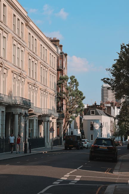 A quiet residential street in Kentish Town with a row of elegant multi-storey terraced buildings featuring cream-colored facades, decorative window frames, and balconies, set against a blue sky with some scattered clouds. The street has parked cars along the curb, including a dark vehicle and a white van, with some pedestrians visible on the pavement. In the foreground, there are stacking cardboard boxes and wrapped furniture, indicating an ongoing house move or home relocation process. The surroundings include trees providing greenery and partial views of modern high-rise buildings in the background. This scene reflects urban moving and packing activities, aligning with services offered by Man with Van Kentish Town for removals and furniture transport.