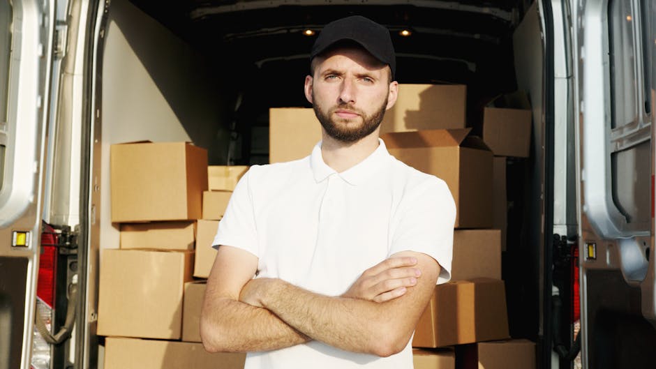 A man with a beard, wearing a white shirt and black cap, stands inside the rear of a moving van, holding a cardboard box during a home relocation process. The van is packed with numerous cardboard boxes of various sizes, some stacked on top of each other, and secured with plastic wrap and packing materials. The loading area inside the van is visible, with a floor designed for transporting household belongings. Outside the van, part of a roadside and residential buildings are visible, suggesting the loading is occurring in a residential neighbourhood. The lighting is natural, indicating daylight. The scene illustrates furniture transport and packing during a same-day moving service, with the van positioned on a street, and the individual preparing items for unloading or organization, consistent with professional removal services by Man with Van Kentish Town.